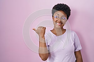 Young african american woman standing over pink background smiling with happy face looking and pointing to the side with thumb up