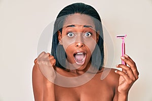 Young african american woman standing topless holding razor screaming proud, celebrating victory and success very excited with