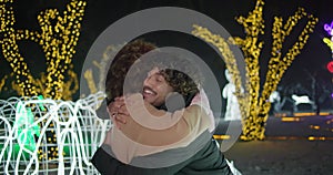 Young African American woman surprising Indian man closing his eyes with arms standing on garlands and lights background