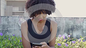 Young African American woman using a mobile phone and sitting on a bench in the park. Female black jogger resting and