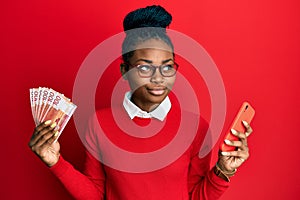 Young african american woman using smartphone holding norwegian krone banknotes smiling looking to the side and staring away