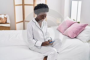 Young african american woman using smartphone sitting on bed at bedroom