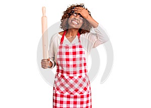 Young african american woman wearing apron holding rolling pin stressed and frustrated with hand on head, surprised and angry face