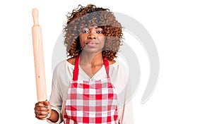 Young african american woman wearing apron holding rolling pin thinking attitude and sober expression looking self confident