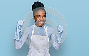 Young african american woman wearing apron screaming proud, celebrating victory and success very excited with raised arms