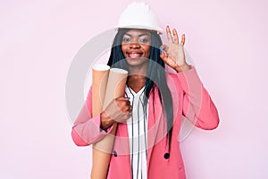 Young african american woman wearing architect hardhat holding blueprints doing ok sign with fingers, smiling friendly gesturing