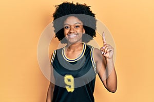 Young african american woman wearing basketball uniform showing and pointing up with finger number one while smiling confident and