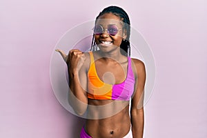 Young african american woman wearing bikini and sunglasses smiling with happy face looking and pointing to the side with thumb up
