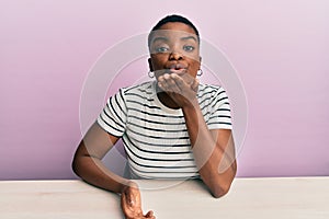 Young african american woman wearing casual clothes sitting on the table looking at the camera blowing a kiss with hand on air