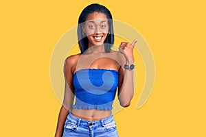 Young african american woman wearing casual summer clothes smiling with happy face looking and pointing to the side with thumb up