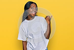 Young african american woman wearing casual white t shirt smiling with happy face looking and pointing to the side with thumb up