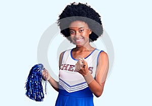 Young african american woman wearing cheerleader uniform holding pompom beckoning come here gesture with hand inviting welcoming