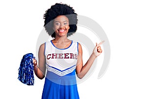 Young african american woman wearing cheerleader uniform holding pompom cheerful with a smile on face pointing with hand and