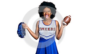 Young african american woman wearing cheerleader uniform holding pompom and football ball sticking tongue out happy with funny