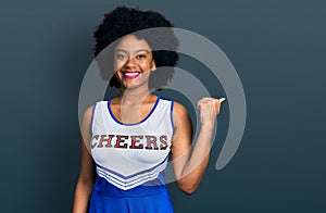 Young african american woman wearing cheerleader uniform smiling with happy face looking and pointing to the side with thumb up