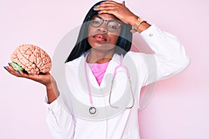 Young african american woman wearing doctor coat holding brain stressed and frustrated with hand on head, surprised and angry face
