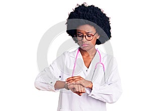 Young african american woman wearing doctor coat and stethoscope checking the time on wrist watch, relaxed and confident