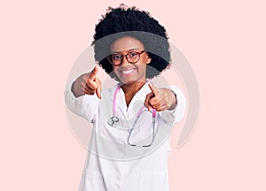 Young african american woman wearing doctor coat and stethoscope pointing to you and the camera with fingers, smiling positive and