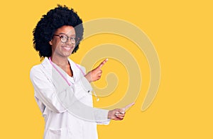 Young african american woman wearing doctor coat and stethoscope smiling and looking at the camera pointing with two hands and