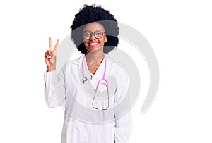 Young african american woman wearing doctor coat and stethoscope smiling looking to the camera showing fingers doing victory sign