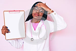 Young african american woman wearing doctor stethoscope holding clipboard stressed and frustrated with hand on head, surprised and