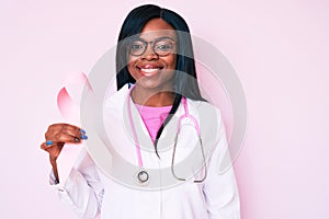 Young african american woman wearing doctor stethoscope holding pink cancer ribbon looking positive and happy standing and smiling