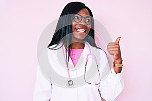 Young african american woman wearing doctor stethoscope smiling with happy face looking and pointing to the side with thumb up