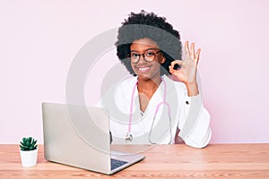 Young african american woman wearing doctor stethoscope working using computer laptop doing ok sign with fingers, smiling friendly