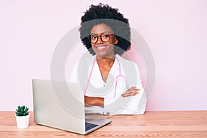 Young african american woman wearing doctor stethoscope working using computer laptop happy face smiling with crossed arms looking