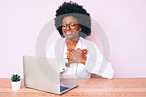 Young african american woman wearing doctor stethoscope working using computer laptop smiling with hands on chest with closed eyes