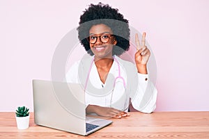 Young african american woman wearing doctor stethoscope working using computer laptop smiling with happy face winking at the
