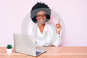 Young african american woman wearing doctor stethoscope working using computer laptop smiling happy pointing with hand and finger