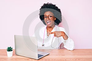 Young african american woman wearing doctor stethoscope working using computer laptop smiling happy pointing with hand and finger
