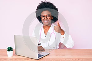 Young african american woman wearing doctor stethoscope working using computer laptop smiling happy and positive, thumb up doing