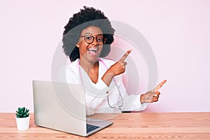 Young african american woman wearing doctor stethoscope working using computer laptop smiling and looking at the camera pointing