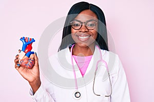 Young african american woman wearing doctor uniform holding heart looking positive and happy standing and smiling with a confident