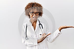 Young african american woman wearing doctor uniform and stethoscope amazed and smiling to the camera while presenting with hand