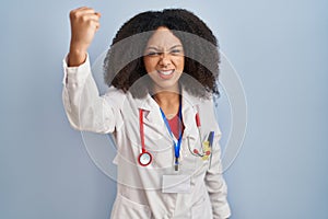 Young african american woman wearing doctor uniform and stethoscope angry and mad raising fist frustrated and furious while
