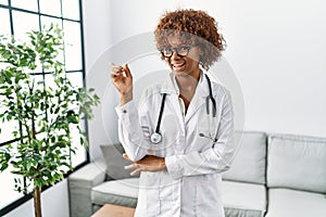 Young african american woman wearing doctor uniform and stethoscope with a big smile on face, pointing with hand and finger to the