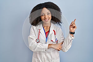 Young african american woman wearing doctor uniform and stethoscope with a big smile on face, pointing with hand finger to the