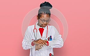 Young african american woman wearing doctor uniform and stethoscope checking the time on wrist watch, relaxed and confident