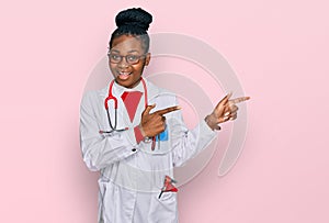 Young african american woman wearing doctor uniform and stethoscope smiling and looking at the camera pointing with two hands and