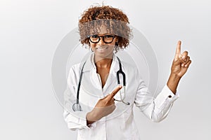 Young african american woman wearing doctor uniform and stethoscope smiling and looking at the camera pointing with two hands and