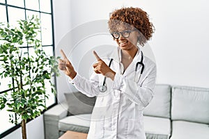 Young african american woman wearing doctor uniform and stethoscope smiling and looking at the camera pointing with two hands and