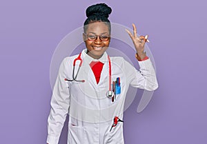 Young african american woman wearing doctor uniform and stethoscope smiling looking to the camera showing fingers doing victory