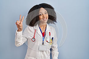 Young african american woman wearing doctor uniform and stethoscope smiling looking to the camera showing fingers doing victory