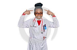 Young african american woman wearing doctor uniform and stethoscope smiling pointing to head with both hands finger, great idea or