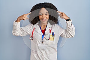 Young african american woman wearing doctor uniform and stethoscope smiling pointing to head with both hands finger, great idea or