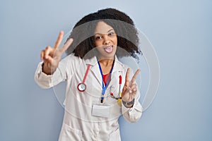 Young african american woman wearing doctor uniform and stethoscope smiling with tongue out showing fingers of both hands doing