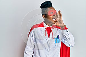 Young african american woman wearing doctor uniform and super hero costume shouting and screaming loud to side with hand on mouth